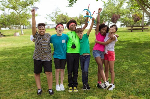 A cheerful group of children playing and celebrating with arms raised at a park on a sunny day.